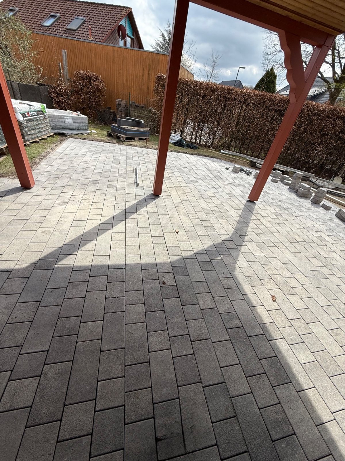 Neu gepflasterte Terrasse unter einem Carport, dahinter eine geschnittene Hecke
